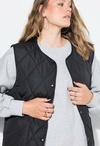 Frau mit welligem Haar, die große Creolen trägt, einen hellgrauen Sweatshirt, eine schwarze gesteppte Weste und mehrere Ringe an beiden Händen.