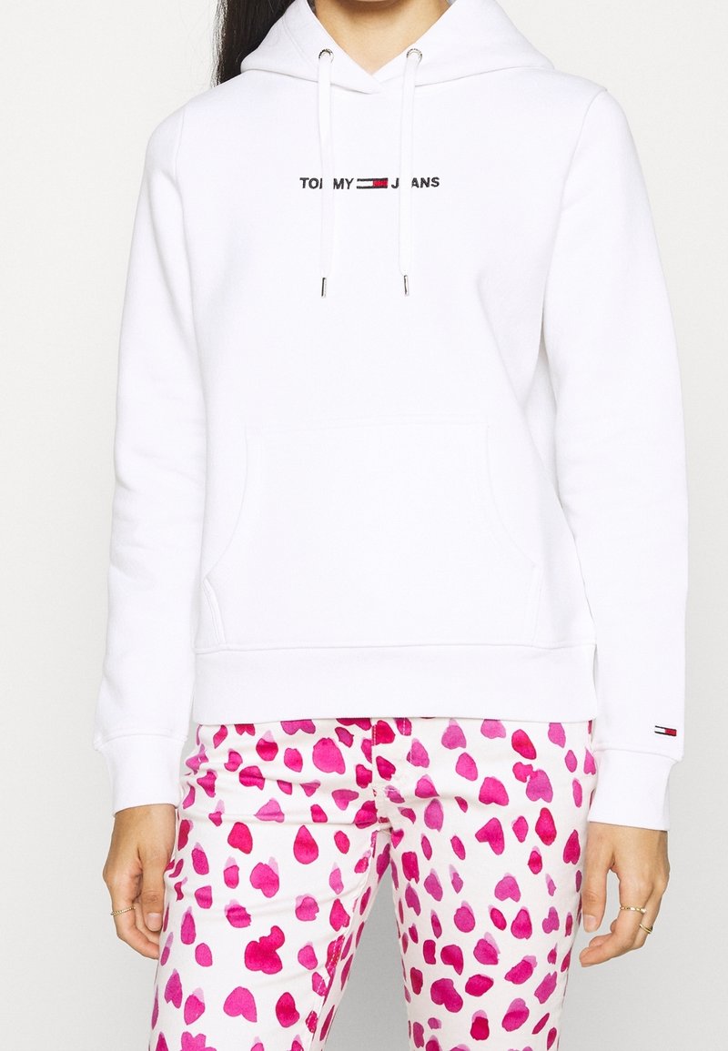 Sudadera blanca con bolsillo frontal y capucha ajustable con cordón, que presenta el logotipo "TOMMY JEANS" bordado en negro. Combinada con pantalones rosa con un patrón de corazones.