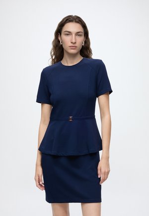 Femme aux longs cheveux bouclés portant un top peplum bleu marine à manches courtes et une jupe assortie sur un fond blanc uni.