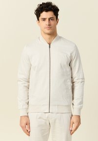 Lichtbeige bomberjack met een ritssluiting aan de voorkant, geribde manchetten en onderkant. Glad materiaal met een minimale textuur en geen zichtbare patronen.