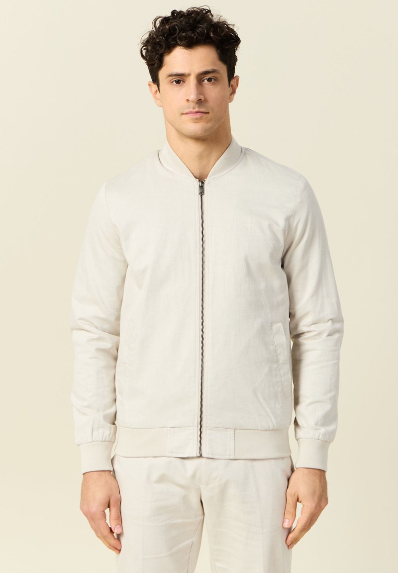 Lichtbeige bomberjack met een ritssluiting aan de voorkant, geribde manchetten en onderkant. Glad materiaal met een minimale textuur en geen zichtbare patronen.