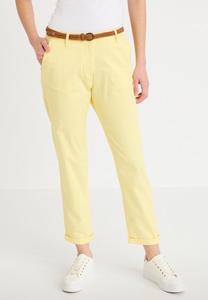 Persoon draagt lichtgele opgerolde broek, bruine gevlochten riem, witte sneakers en een wit shirt, staand met één hand in de zak.