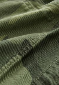 Grön kamouflagematerial med en texturerad väv, som visar distinkta mönster och sömnadsdetaljer, vilket framhäver hållbarhet och materialkvalitet.