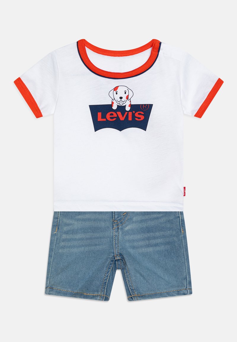 Bijela pamučna majica s crvenim detaljima, koja ima grafiku psa i mornarsko plavi logo. Uparena s plavim trapericama kratkim hlačama s žutim šavovima i džepovima.