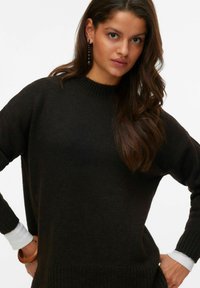 Suéter de punto marrón oscuro con un cuello alto, mangas anchas y puños acanalados. Se ve una camisa blanca en los puños. Textura suave.