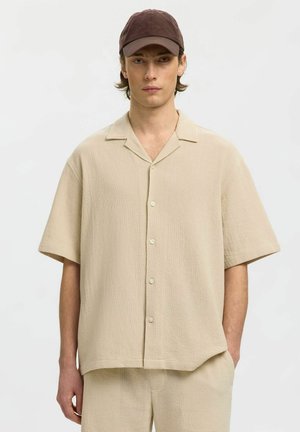 Jeune homme portant une chemise beige à manches courtes boutonnée et un pantalon assorti, avec une casquette marron foncé, debout devant un fond uni.