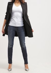 Abrigo negro largo con detalles de botones sobre una camiseta blanca de cuello en V, combinado con jeans ajustados azul oscuro y tacones altos con un patrón plateado.