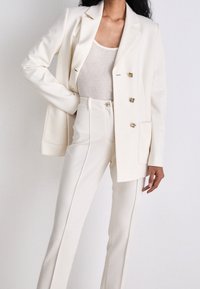 Blazer blanc ajusté avec un devant croisé et des boutons dorés, associé à un pantalon taille haute assorti, fabriqué en tissu lisse.
