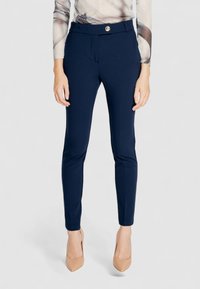 Pantalons ajustés bleu marine, avec une coupe slim, dotés d'une fermeture à bouton frontal et d'une texture lisse. Portés avec des chaussures à talons hauts couleur nude.