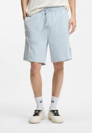 Hombre con pantalones cortos azul claro con cordón, calcetines blancos con logotipos azules y zapatillas blancas con cordones negros, de pie frente a un fondo blanco.