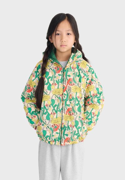 Kind met lang gevlochten haar, gekleed in een kleurrijk, bloemenpatroon capuchonjack en lichtgrijze broek, staand met handen in de zakken.