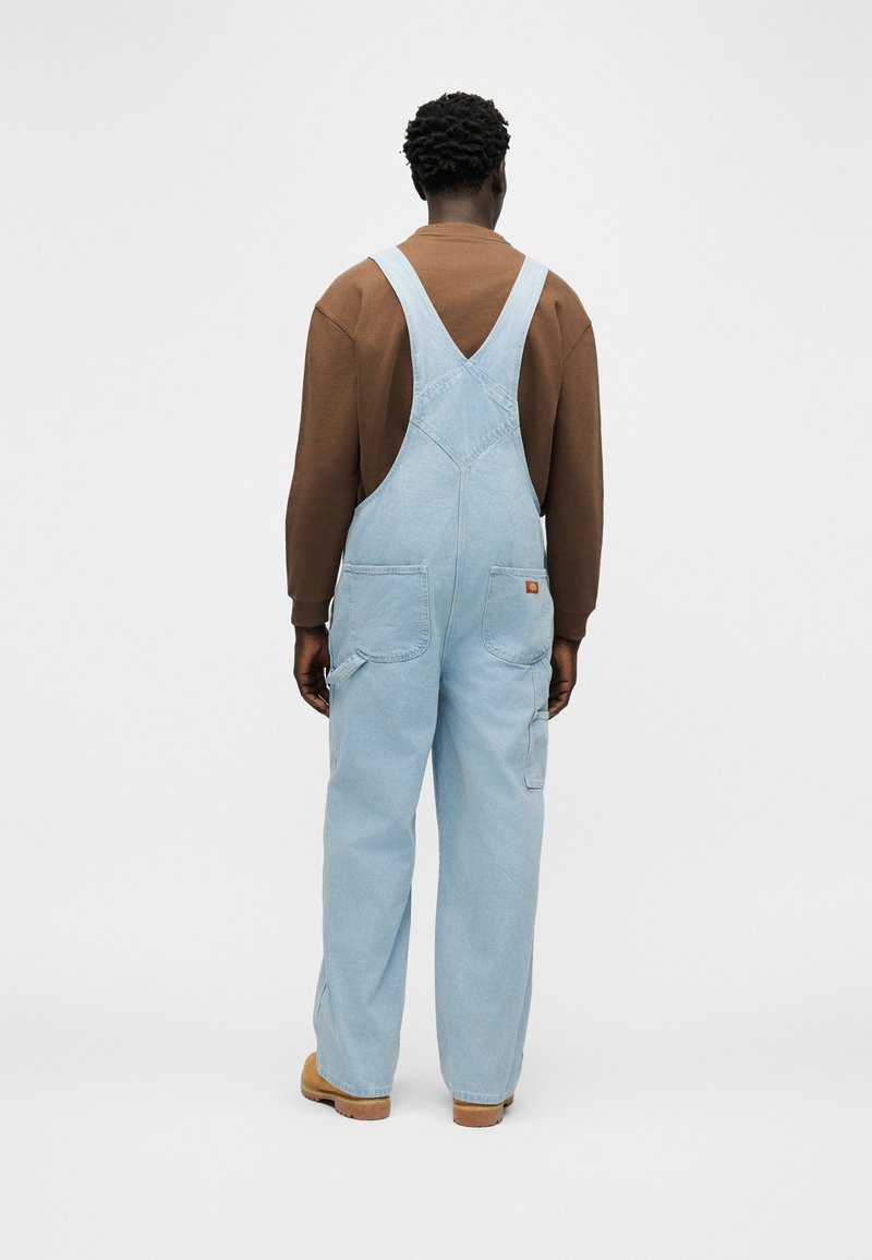 Person trägt hellblaue Jeans-Latzhose und braunes langärmliges Hemd, blickt weg, steht vor einem schlichten weißen Hintergrund.