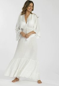 Vestido largo blanco con un escote en V profundo y cintura con cordón. Presenta mangas amplias y fluidas, y un dobladillo con volantes en capas. La tela tiene un brillo sutil.