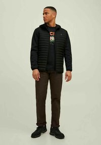 Ung mand står op, ser til siden, iført en sort quilted jakke med hætte, en trykt sort sweatshirt, brune bukser og sorte sko.