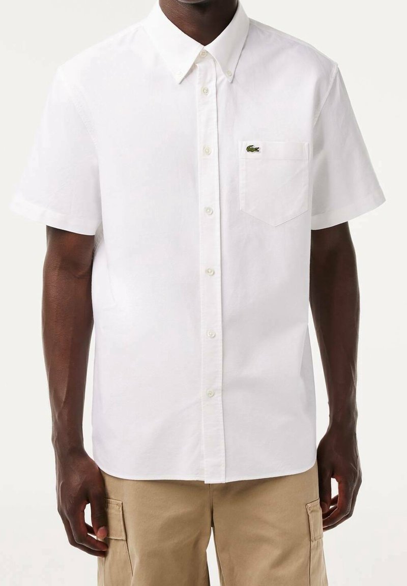Homme portant une chemise blanche à manches courtes avec une poche poitrine et un pantalon cargo beige sur un fond uni.