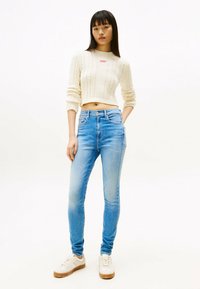Suéter de punto acanalado color crema con un diseño recortado y un logo rojo. Jeans ajustados azul claro con un lavado deslavado. Zapatillas blancas con suelas marrones.