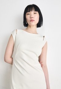 Form-fitting, sleeveless dress in soft ivory fabric. Features cut-out shoulder detail and smooth, draped texture.
