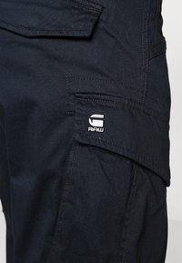 Temno modri cargo hlače iz strukturirane tkanine z bočno žepom in majhnim belim logotipom, poudarjene z vidnimi šivi.