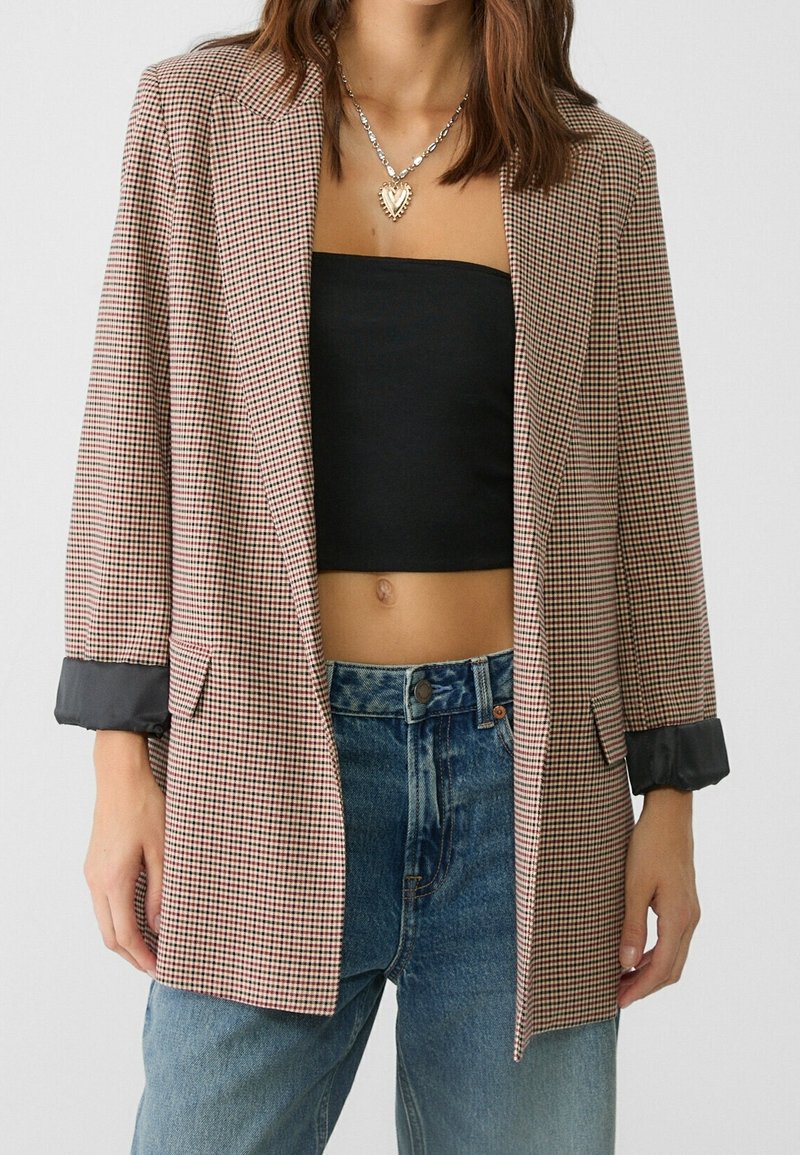 Blazer a cuadros en tejido marrón y crema, con un corte relajado, solapas con muesca y puños negros en contraste. Llevado sobre una blusa negra y vaqueros.