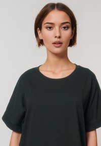 Een jonge vrouw met kort, bruin haar draagt een donker T-shirt en kijkt recht in de camera met een neutrale uitdrukking tegen een lichte achtergrond.