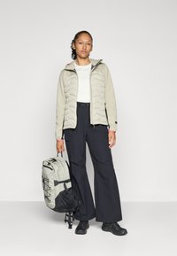 Beige paksu takki hupulla, valkoinen pitkähiha, mustat leveät housut ja harmaa reppu mustilla yksityiskohdilla ja hihnoilla.