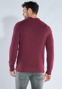 Gerippter bordeauxroter Pullover mit hohem Kragen, figurbetontem Schnitt und langen Ärmeln, kombiniert mit grauer Jeans mit Gesäßtaschen.