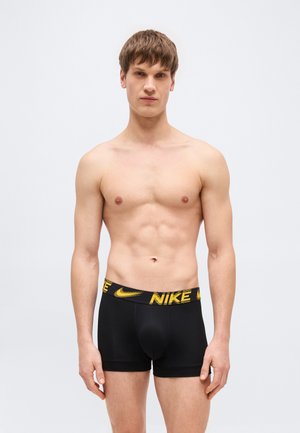 Noor mees seisab, seljas mustad Nike bokserid kollase logoga vöökohal, heleda tausta ees.