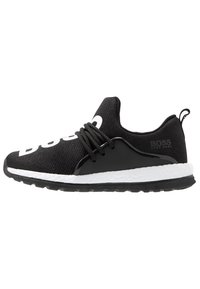 Zapatilla BOSS negra con suela gruesa blanca, punteras redondeadas, cordones negros y la marca "BOSS" en el lado y en la parte superior del tejido.