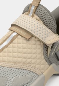 Αθλητικό παπούτσι σε μπεζ και γκρι χρώμα με υφή στο επάνω μέρος που έχει κεντήματα, ανοιχτό γκρι δίχτυ και ιμάντα με βέλκρο με λογότυπο.