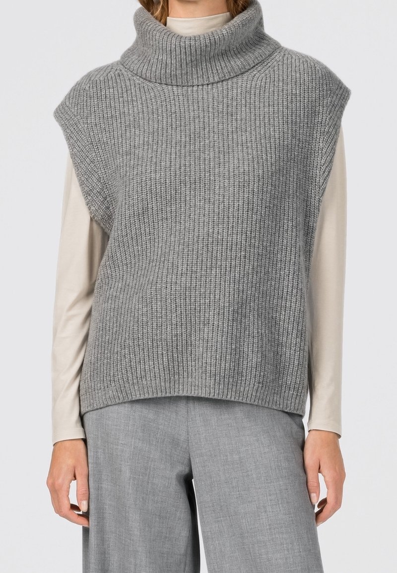 Gilet sans manches en tricot gris épais avec col roulé porté sur un top beige à manches longues et un pantalon gris à jambes larges.