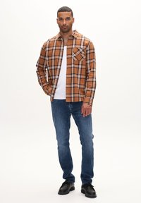 Brun ternet skjorte med sorte og hvide accenter, parret med blå jeans og sorte sko. Skjorten har en enkelt brystlomme.