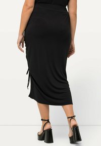 Jupe midi noire en tissu doux, avec des attaches latérales et un design légèrement ajusté. Le modèle porte des chaussures à talons aiguilles noires.