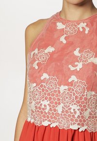 Korálové šaty s průhledným výšivkovým krajkovým tělem se květinovými vzory a šestiúhelníkovým designem. Spodní část má volný sukňový vzhled.