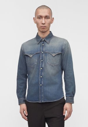 Homme aux cheveux courts portant une chemise en jean bleue délavée à boutons avec poches poitrine et un pantalon noir, debout devant un fond clair uni.