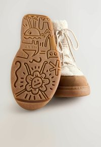Hvide støvler med fleece-for, der har en tan suede tå og gummisul. Sålen har intrikate præget grafik, herunder en smilende blomst og forskellige former.