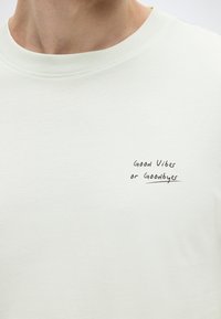 Ανοιχτά πράσινο βαμβακερό μπλουζάκι με στρογγυλή λαιμόκοψη και κοντά μανίκια, με μαύρο χειρόγραφο κείμενο που γράφει "Καλοί ρυθμοί ή αποχαιρετισμοί."