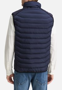 Veste matelassée sans manches bleu marine avec quilting horizontal, col montant et panneaux latéraux contrastants. Portée avec une chemise blanche à manches longues et un jean.