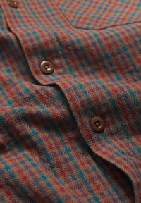 Gestructureerd shirt met een levendig patroon van rood, blauw en paars tegen een bruine achtergrond, met bruine knopen en een gladde afwerking.