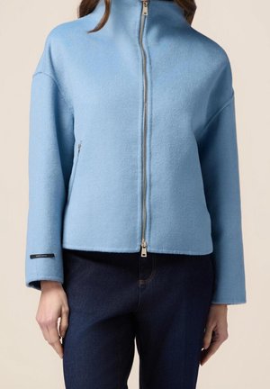 Veste bleu clair zippée avec col montant portée sur un jean bleu foncé, montrée de la moitié du torse jusqu'aux poignets.