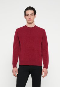 Punane kootud kampsun tekstuurse, mustrilise disainiga, soonilise ümarakaeluse ja pikkade varrukatega. Kandatud mustade pükstega neutraalse tausta ees.