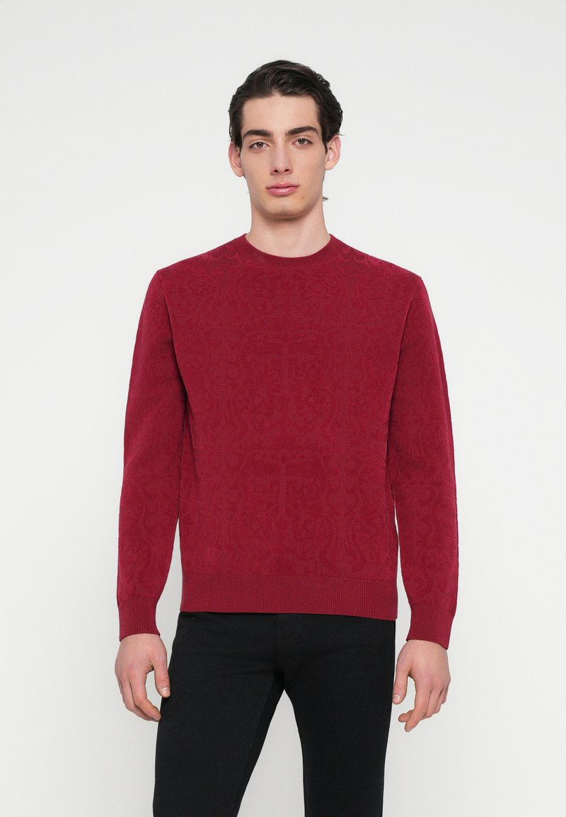 Punane kootud kampsun tekstuurse, mustrilise disainiga, soonilise ümarakaeluse ja pikkade varrukatega. Kandatud mustade pükstega neutraalse tausta ees.