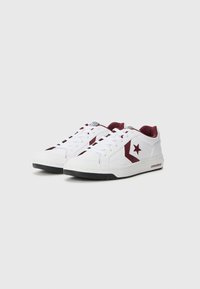 White sneakers with burgundy accents, featuring textured leather, rounded toe, lace-up closure, and a logo star on the outer side.
