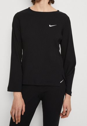 Personne portant un haut noir côtelé à manches longues et un pantalon assorti avec des logos blancs Nike swoosh sur la poitrine et la hanche.