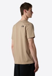 Beige bomulds T-shirt med rund hals, korte ærmer og et lille sort logo på den øverste del af ryggen. Glat tekstur, afslappet pasform.
