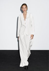 Donna che cammina, indossando un abito bianco ampio con pantaloni larghi e zoccoli neri, su uno sfondo bianco semplice e con pavimento scuro.