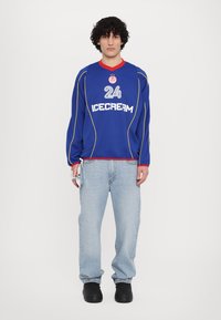 Blauw langemouwen shirt met gele accenten, witte tekst "ICECREAM" en het nummer "24." Gecombineerd met lichtblauwe jeans en zwarte schoenen.