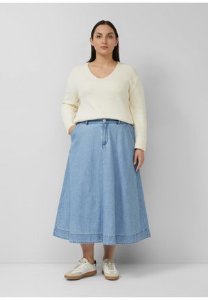 Kobieta stoi z rękami w kieszeniach, ubrana w kremowy prążkowany sweter z dekoltem w serek, niebieską midi spódnicę dżinsową i białe sportowe buty z gumową podeszwą.
