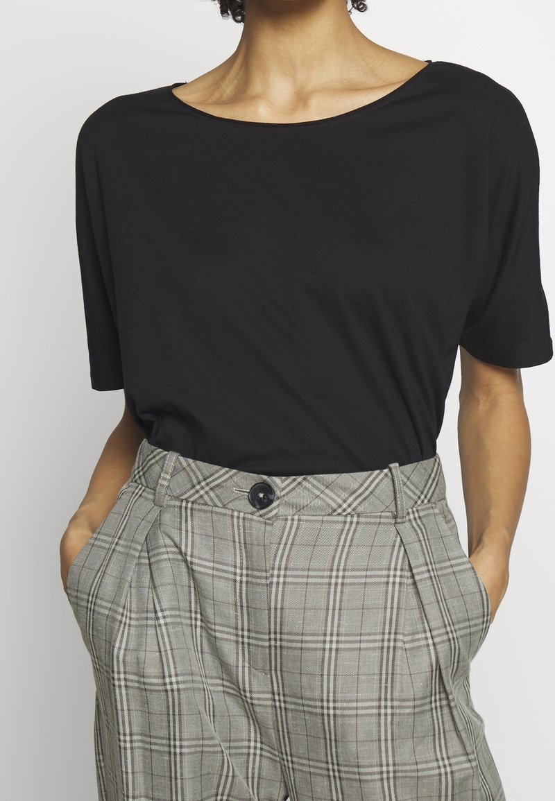 Personne portant un haut noir ample à manches courtes rentré dans un pantalon à carreaux gris taille haute, mains dans les poches.