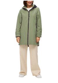 Veste imperméable verte avec capuche noire, fermeture éclair complète et poches latérales. Portée par-dessus un pantalon beige à jambes larges et des baskets blanches.