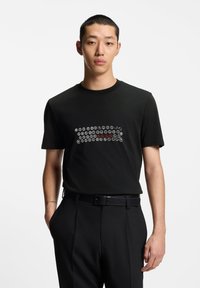 Crna pamučna majica s prednjim grafičkim prikazom bijelih i crvenih kružnih uzoraka. Kratki rukavi, okrugli izrez i opušteni kroj.